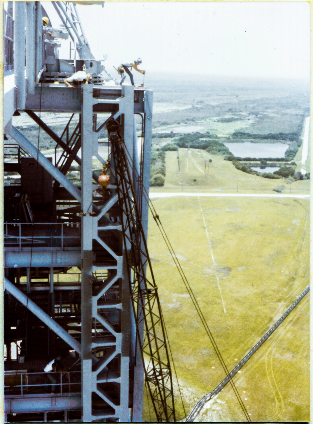 Image 083. Union Ironworker James Dixon has placed a loop of wire rope around the top of the GOX Arm Hinges Support Strongback, and is standing on top of the Fixed Service Structure at Space Shuttle Launch Complex 39-B, Kennedy Space Center, Florida, at Elevation 300'-0”, no handrails, wearing no safety harness or other gear, as was common practice in the early 1980's, working the come-along that the wire rope has been threaded to, via an unseen snatch block which is hidden behind the Strongback. Main force is now being applied, and the top of the Strongback is in hard contact with the web of the Perimeter Framing Beam of the FSS, scraping along against it as it is urged ever northward by the action of the come-along, having been shoved into that hard contact by the further action of other Union Ironworkers two levels down, at Elevation 260'-0”, who are slowly tensioning the wire rope around the second-to-bottom horizontal bracing member at the lower end of the Strongback, forcing it into a more-vertical orientation, going against its natural tendency to hang at an angle because of where its lifting sling remains attached, roughly one-third down from its top. The Strongback is not yet fully vertical and is only contacting the FSS at its top connection point. Below that, the other two connection points remain out of contact, and the Strongback is still reasonably free to be pushed side-to-side, along the face of the FSS. At top and bottom, other Union Ironworkers are in constant communication with the rest of the gang, closely-monitoring the position of the Strongback and its connection points. Photo by James MacLaren.